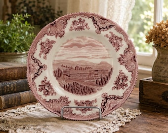 May include: A round ceramic plate featuring a deep red transferware pattern on a cream background. The center depicts a landscape with buildings and trees, while the wide rim is decorated with intricate floral motifs and scrollwork borders.