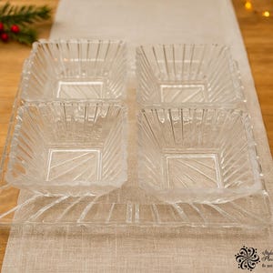 May include: A set of four clear, square glass serving dishes with a textured, ridged design. The dishes are arranged on a clear tray, ideal for serving snacks or appetizers. The image is set on a wooden table with festive decorations.