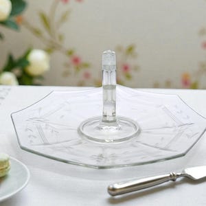 May include: Clear glass serving tray with a handle. The tray has an octagonal shape with etched floral designs. A silver cake server and a plate with macarons are in the foreground. The background is a floral wallpaper.