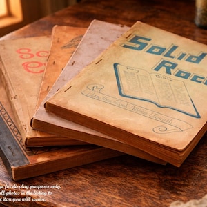May include: A stack of vintage books with aged, tan covers. The top book features the title "Solid Rock" in blue lettering, with an illustration of an open Bible. Other books have faded titles and red lettering. The books are arranged on a wooden surface.