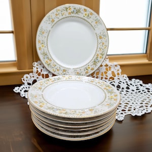 May include: A set of six white ceramic plates with a floral pattern in shades of yellow, green, and blue. The plates have a gold rim and are stacked on top of each other.