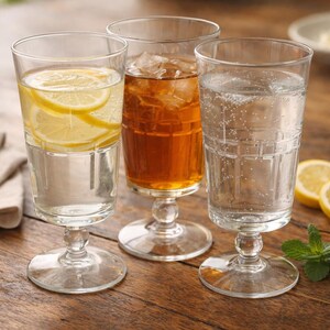 May include: Three stemmed glasses filled with beverages, including water with lemon slices, iced tea, and sparkling water. The glasses sit on a wooden table, with lemon slices and mint sprigs nearby.