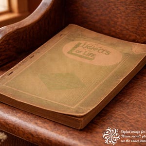 May include: An antique book titled "Lights of Life" rests on a wooden surface. The book has a worn, aged appearance with a tan cover and green accents. The book is placed on a dark brown wooden surface, suggesting a vintage or historical theme.