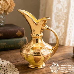 May include: A gold-colored, vintage-style pitcher with a handle and a unique spout design. The pitcher has a textured surface and sits on a wooden table. The pitcher is next to a stack of books and a lace doily.