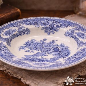 May include: A white and blue ceramic bowl with a floral design. The bowl has a wide rim with a detailed blue pattern and a central blue landscape scene. The bowl is placed on a beige linen cloth.