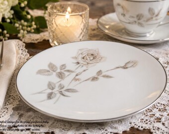 May include: A round white porcelain bread plate featuring a delicate grey rose illustration with leaves and a bud, finished with a thin silver rim. The plate is smooth and circular, showcasing a detailed botanical drawing in the center.