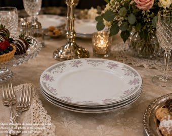 May include: A stack of white porcelain dinner plates featuring a delicate floral pattern in shades of pink and grey along the rim. The plates are circular with a smooth finish and are displayed on a table set with silverware and glassware.
