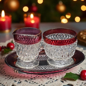 May include: A crystal creamer and cup set with a diamond pattern and a red rim, resting on a matching silver-toned tray. The set is displayed on a lace tablecloth, with lit candles and Christmas decorations in the background.
