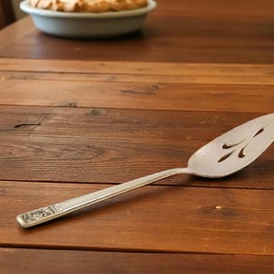 May include: A silver pie server with a decorative handle and a slotted blade. The server is resting on a wooden table. The blade has a decorative cut-out design.