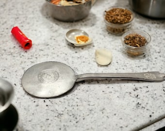 May include: A silver metal spoon with a round, flat head and a handle. The head of the spoon has a circular design with the letters "AK" in the center. The spoon is lying on a white countertop.