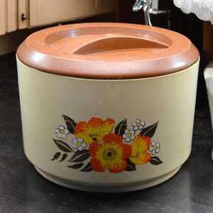 May include: A vintage, cream-colored, round plastic container with a brown lid. The container has a floral design with orange, yellow, and white flowers.