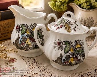 May include: A white ceramic creamer and a lidded sugar bowl featuring a colorful transferware pattern of a rural landscape with a yellow house, green trees, and red berries. Both pieces have a scalloped rim and curved handles with a glossy finish.