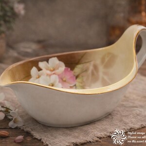 May include: Cream-colored gravy boat with a gold rim and handle. The interior features a floral design with pink and white flowers. The boat is set on a linen cloth with a biscuit and flower petals.