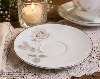 May include: A white porcelain saucer featuring a delicate gray rose illustration with leaves and a thin silver rim. The saucer has a central circular indentation for a cup and a smooth, glossy finish, presenting a clean and refined appearance.