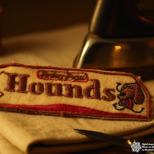 May include: Embroidered patch with the word "Hounds" in brown thread on a cream-colored background. The patch features a cartoon dog illustration and red border. A vintage sewing machine and scissors are in the background.