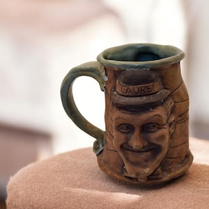May include: A brown ceramic mug with a sculpted face and the word "LAUREL" on the hat. The mug has a green glaze on the rim and handle.