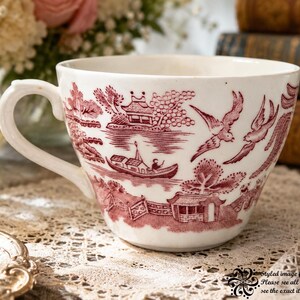 May include: A white ceramic teacup featuring a detailed maroon transferware pattern depicting a landscape with pagodas, a boat on water, and flying birds. The handle is curved and the cup has a smooth, rounded rim and a wide, tapered base.