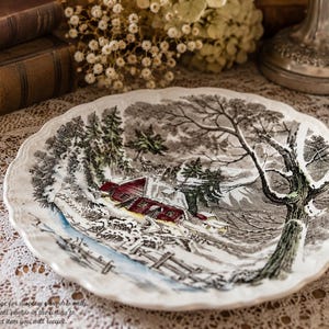 May include: A round ceramic plate with a scalloped edge featuring a detailed winter scene. The illustration depicts a red barn surrounded by snow-covered ground, evergreen trees, and a large bare tree, all rendered in shades of black, white, and red.