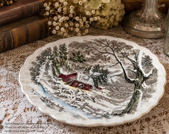 May include: A round ceramic plate with a scalloped edge featuring a detailed winter scene. The illustration depicts a red barn surrounded by snow-covered ground, evergreen trees, and a large bare tree, all rendered in shades of black, white, and red.