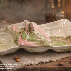 May include: A vintage divided serving dish with a lid. The dish is off-white with a marbled design in shades of pink and green. The lid has a handle. The dish is on a wooden surface with almonds and crackers.
