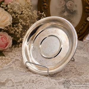 May include: A round silver-toned saucer with a smooth, reflective surface and a raised central circular indentation. The saucer is displayed upright on a thin, silver-colored metal wire stand, showcasing its metallic finish and circular rim.