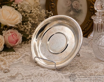 May include: A round silver-toned saucer with a smooth, reflective surface and a raised central circular indentation. The saucer is displayed upright on a thin, silver-colored metal wire stand, showcasing its metallic finish and circular rim.