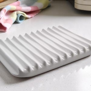 May include: A white ceramic soap dish with a ridged design. The rectangular dish has a series of parallel, curved ridges across its surface. The dish is clean and modern, suitable for a bathroom or kitchen setting.