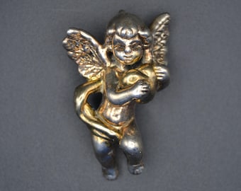 May include: A silver and gold cherub figurine holding a heart. The cherub has wings and is standing on a base.