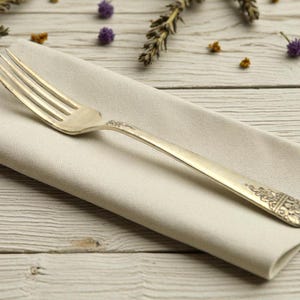 May include: A silver fork with an ornate handle rests on a beige linen napkin. The fork has four tines and a detailed design on the handle. The napkin is folded and placed on a light-colored wooden surface, with sprigs of lavender and small purple flowers.
