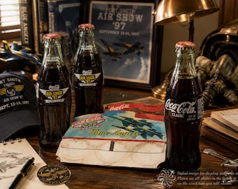 May include: Three glass Coca-Cola bottles, two featuring Fort Smith Air Show '97 labels and one labeled Coca-Cola Classic 8 fl oz. The bottles are accompanied by a cardboard carrier decorated with Blue Angels aircraft graphics and text.