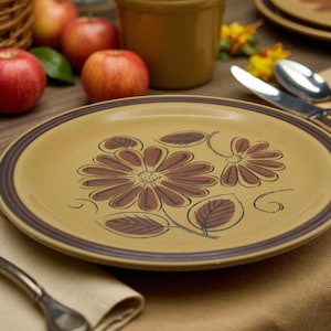 May include: A round, tan-colored plate with a brown floral design and a dark brown border. The plate is part of a dinnerware set, with a knife and fork on a beige napkin. Apples and a basket are in the background.