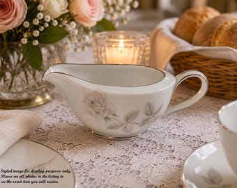 May include: A white porcelain gravy boat featuring a delicate grey rose floral pattern and a silver rim. The vessel has a curved pouring spout and a single handle, designed for serving sauces or cream at a table setting with a refined aesthetic.