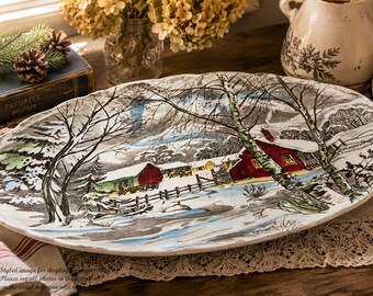 May include: An oval ceramic serving platter featuring a detailed winter landscape illustration. The scene depicts snow-covered ground, bare trees, and two red houses with yellow windows. The color palette includes white, black, red, yellow, and blue.