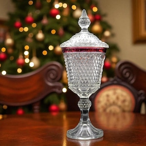 May include: A clear, ornate glass candy dish with a diamond pattern and a red rim. The dish has a lid with a pointed finial and sits on a pedestal base. The background features a Christmas tree with colorful ornaments.
