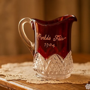May include: A vintage ruby red glass pitcher with a clear glass handle. The words "Worlds Fair 1904" are etched on the front. The bottom of the pitcher has a diamond-cut pattern. It sits on a lace doily.
