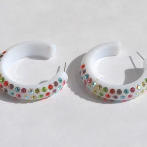 May include: A pair of white hoop earrings featuring a C-shaped design. The outer surface of each hoop is decorated with multiple rows of small, multicolored rhinestones in shades of red, green, blue, and pink, set into the white material.