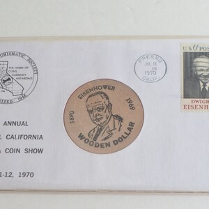 May include: A white envelope with a brown stamp featuring a portrait of Dwight D. Eisenhower. The envelope has a circular stamp with a portrait of Eisenhower and the text "1890 Eisenhower 1969 Wooden Dollar". The envelope also has a circular stamp with the text "Fresno Jul 11 Am 1970 Calif.". The envelope has a circular stamp with the text "Fresno Numismatic Society The Hobby of Coins Currency and Medals Organized 1946". The envelope has the text "4th Annual Central California Stamp & Coin Show July 11-12, 1970".