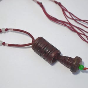 May include: A dark brown pendant necklace with a cylindrical and carved design. The necklace features a red cord, green and white beads, and a small green bead at the bottom. The necklace is displayed on a white surface.