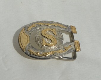 May include: Silver-toned horseshoe belt buckle with gold-colored accents. The buckle features a gold-colored letter "S" in the center, surrounded by a textured silver background and decorative gold flourishes. The buckle is designed for a belt.