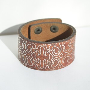May include: A brown leather cuff bracelet with a silver embossed pattern. The bracelet has two silver snaps.