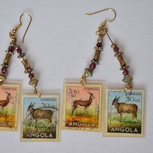 May include: A pair of earrings featuring three different Angolan postage stamps with images of antelopes. The stamps are attached to gold-colored beads and wire.