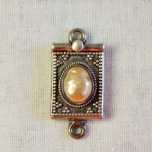 May include: A silver-toned pendant with a pearl-like stone set in a decorative frame. The pendant has two loops for attaching to a chain or cord.