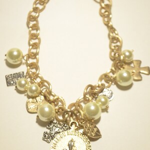 May include: A gold chain necklace with multiple charms, including a fleur-de-lis pendant with the inscription "WAKLOU MES CHERCI YVELINES". The necklace also features faux pearls and other decorative charms.