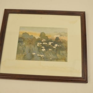 May include: A framed watercolor painting depicting a small village nestled among green trees and hills. The sky is a soft orange and blue, suggesting a sunset or sunrise. The painting is signed "Gail 01".