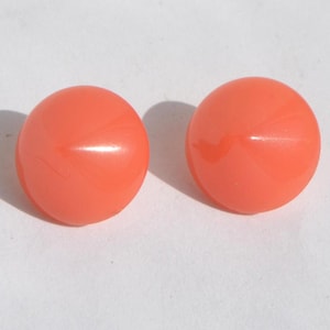 May include: Two round, coral-colored buttons. The buttons are smooth and glossy, with a slight marbling effect visible on their surface. They are set against a white background, with shadows indicating a light source.