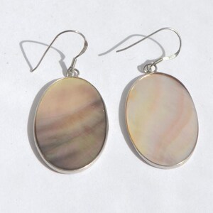 May include: A pair of oval-shaped earrings with a pearlescent, iridescent surface. The earrings have a silver-colored metal border and a hook closure. The colors range from cream to light brown and gray.