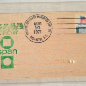 May include: A close-up of a wooden postcard with a green logo that says "span" and a stamp from the American Numismatic Association Convention in Washington, D.C. on August 10, 1971.