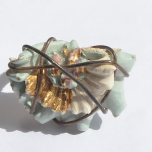 May include: A light blue and gold floral brooch with a wire wrap design. The brooch features gold-colored petals and a light blue base, with a white and pink accent. The wire is a dark brown color.
