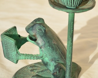 May include: A green metal frog figurine holding a book with a candle holder on top. The frog is sitting on a base with a leaf design.