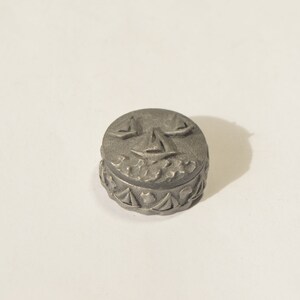 May include: A small, round, silver trinket box with an embossed design of sailboats on the lid. The box has a decorative border around the edge.